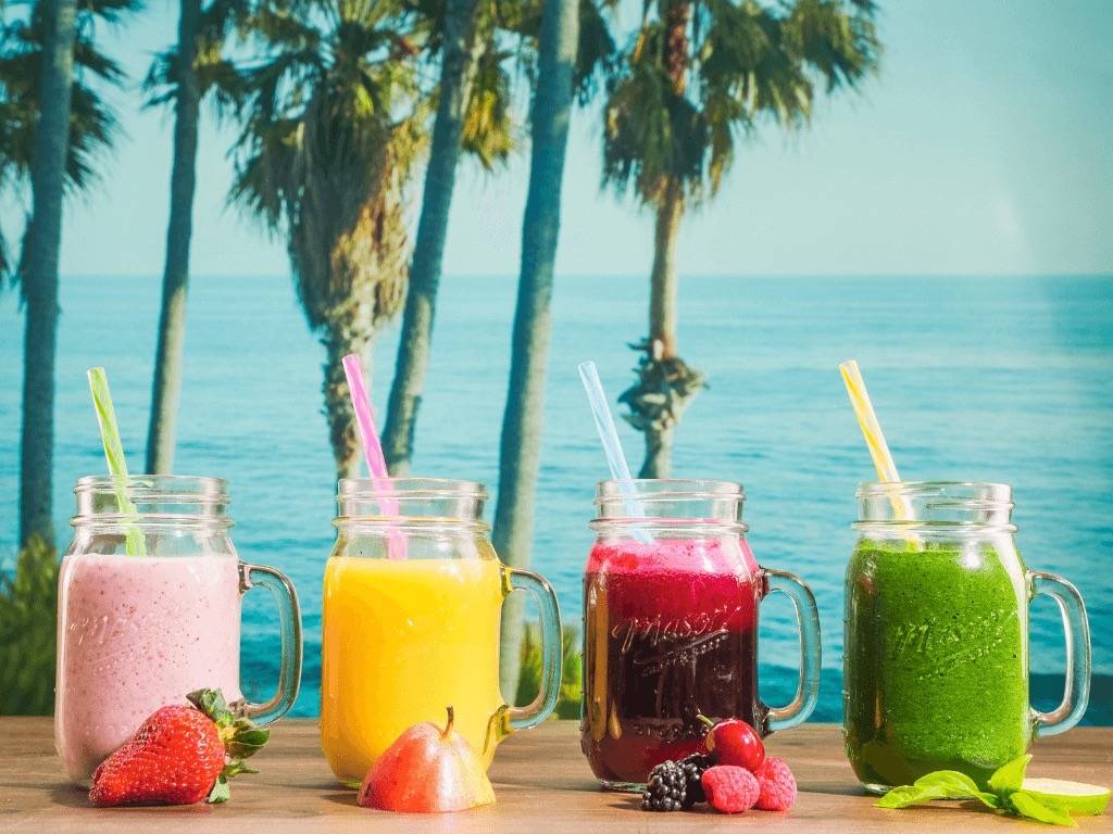 Four colorful detox drinks (strawberry, mango, berry, green) in mason jars by the beach