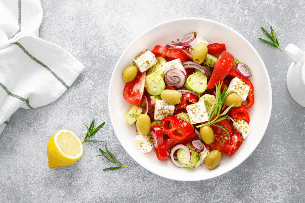 Fresh Greek salad with cucumber, tomatoes, red onion, green olives, and feta cheese cubes served in a white bowl with lemon and rosemary garnish