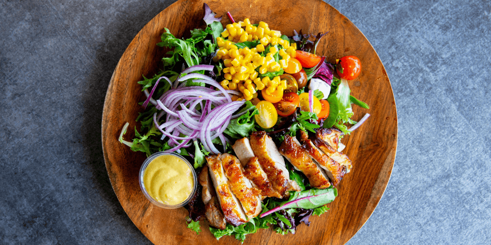 A vibrant plant-based salad with grilled tofu, mixed greens, corn, cherry tomatoes, and red onions, served in a wooden bowl.