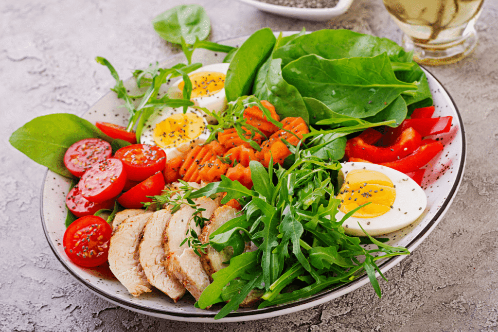 Colorful Mediterranean dinner plate with grilled chicken, spinach, cherry tomatoes, carrots, avocado, and fresh greens in a bowl – healthy and protein-packed