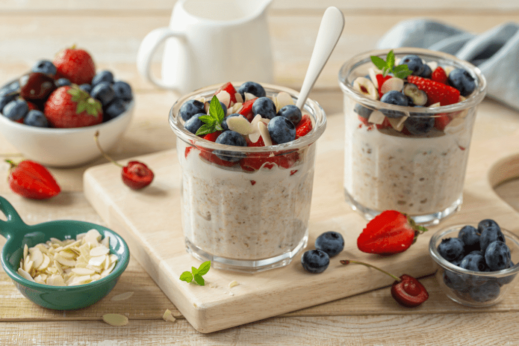 Two glass jars filled with creamy overnight oats, topped with fresh blueberries, strawberries, banana slices, and mint leaves, placed on a wooden board with bowls of fruits and almonds in the background.