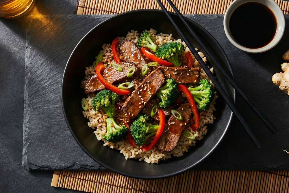 Asian beef and broccoli with brown rice in black ceramic bowl with chopsticks on bamboo mat