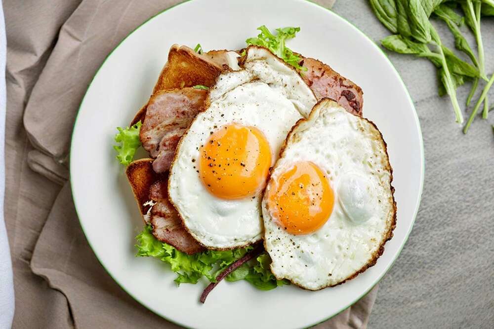 Two fried eggs with bacon and fresh greens on a white plate, high protein breakfast